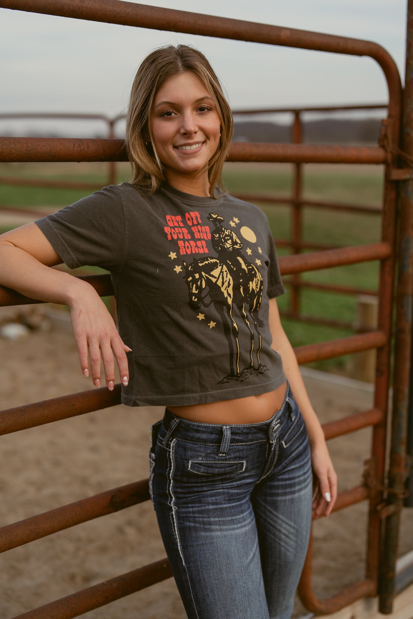 Woman wearing a graphic t-shirt that says get off your high horse and jeans standing next to a rusted metal fence with a field in the background 