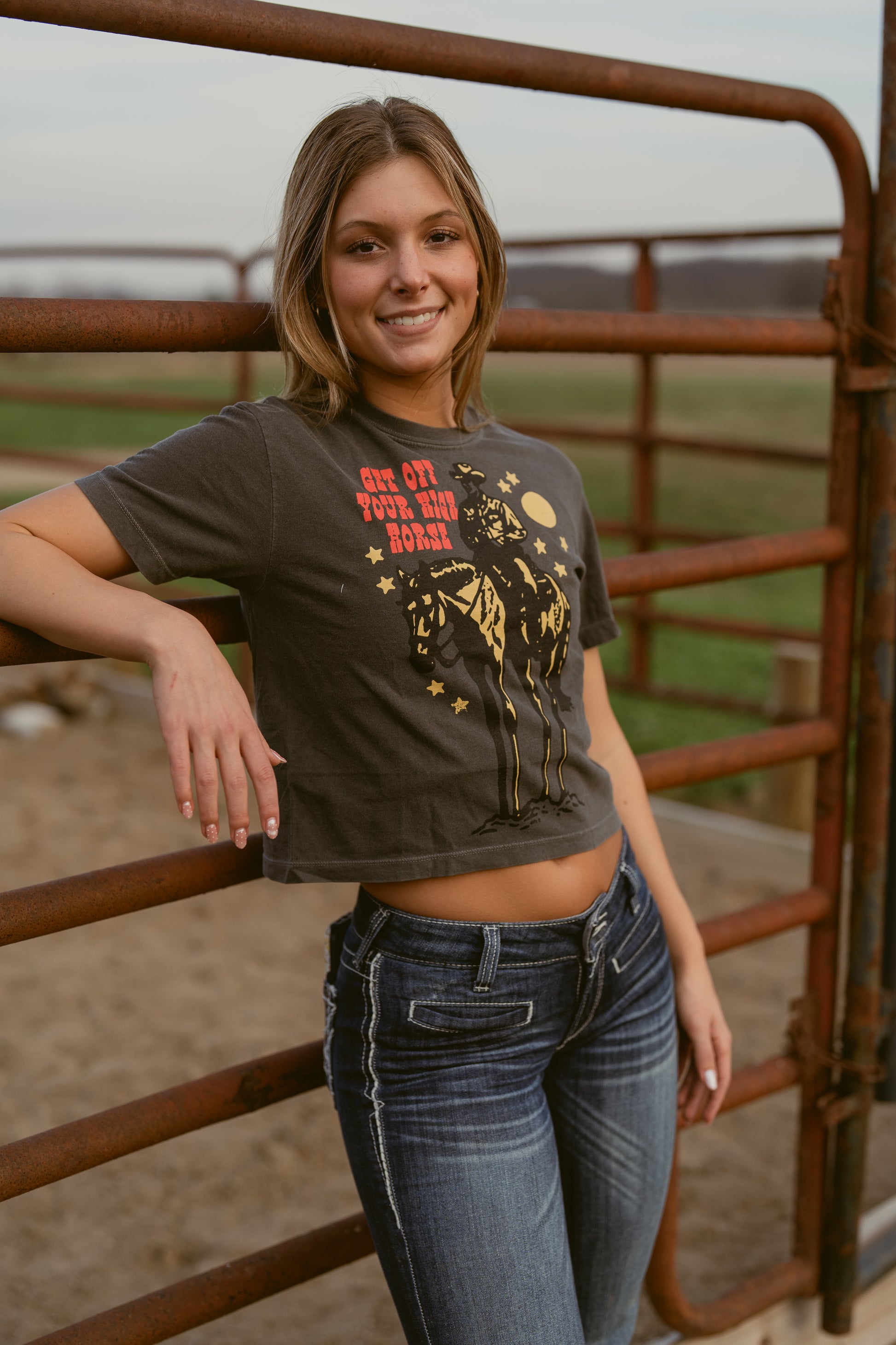 Woman wearing a graphic t-shirt that says get off your high horse and jeans standing next to a rusted metal fence with a field in the background 